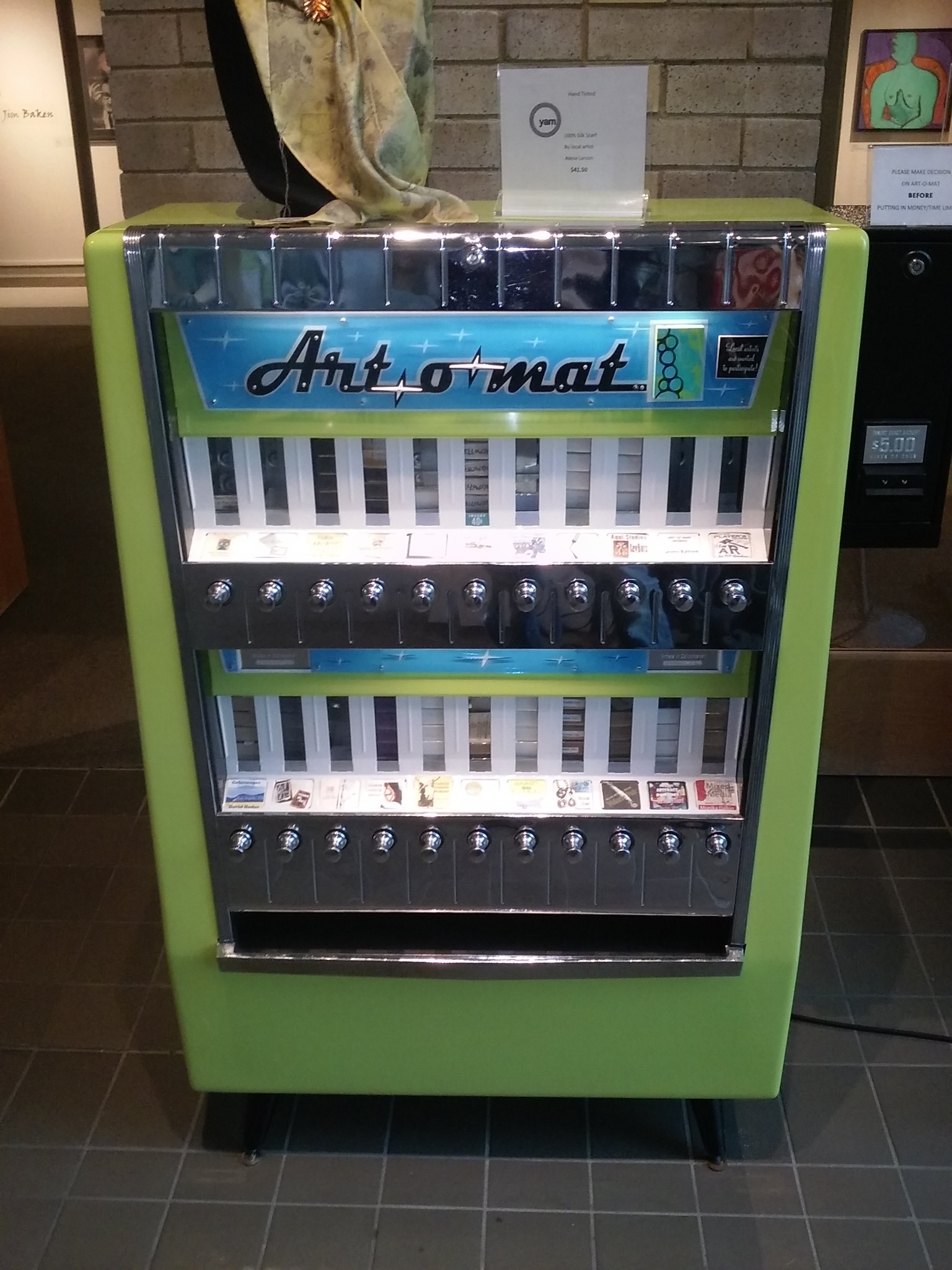 An Art-o-mat machine with a green cabinet, chrome controls and small artworks on display.