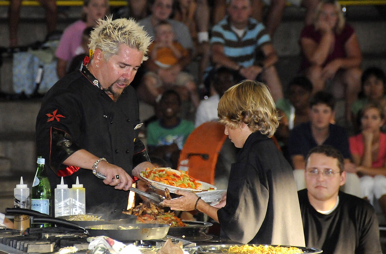 Guy Fieri and his son hold a plate of food during a cooking demonstration, with an audience behind them.