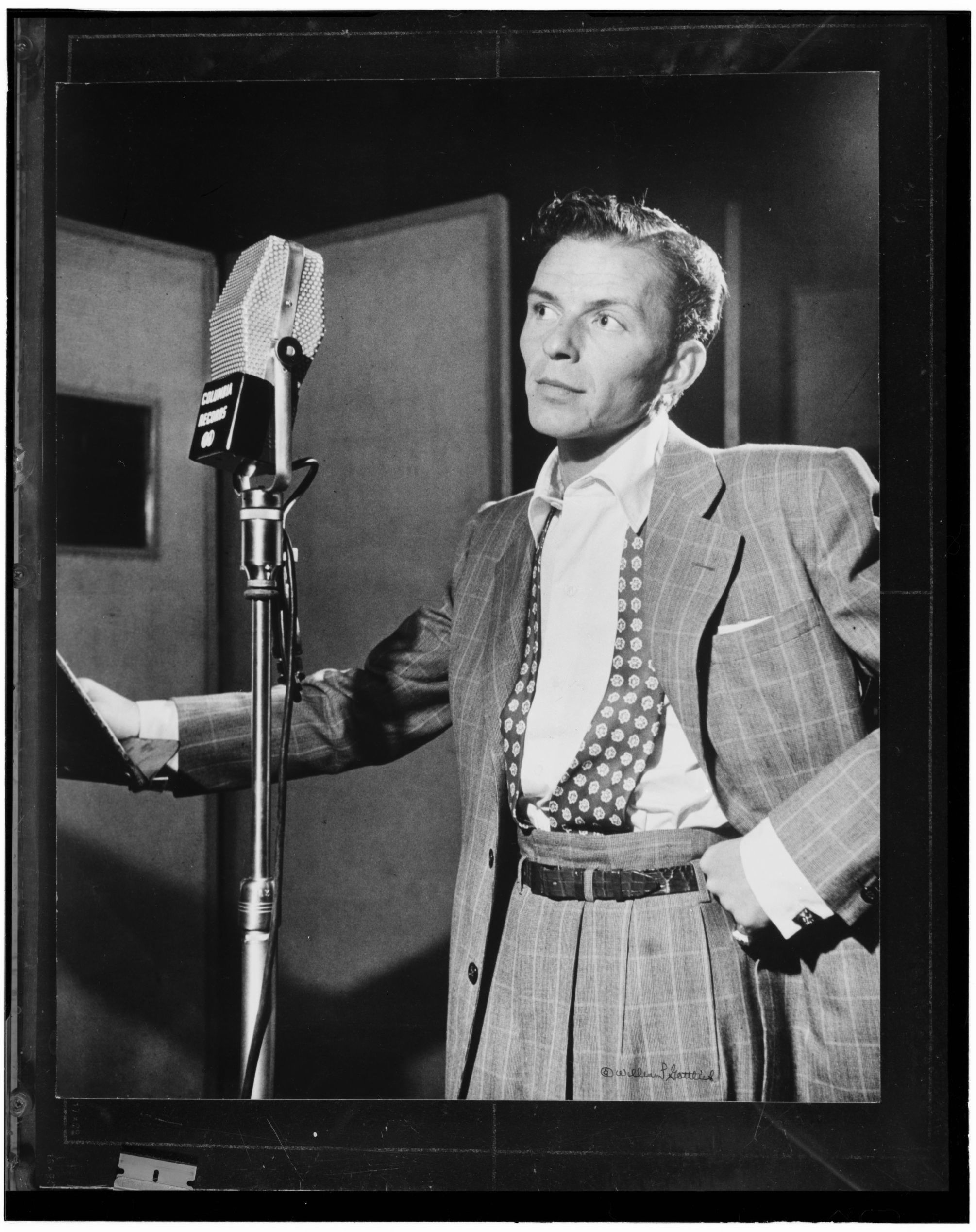 Frank Sinatra in a suit stands beside a studio microphone, holding a sheet of music.