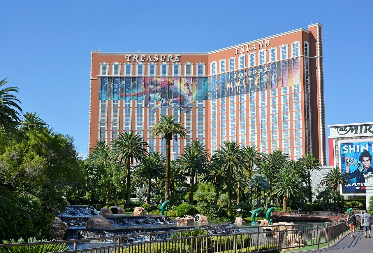 Treasure Island’s peach tower and Mystère mural beyond a terraced waterfall and palms; a Mirage billboard is at the right edge.
