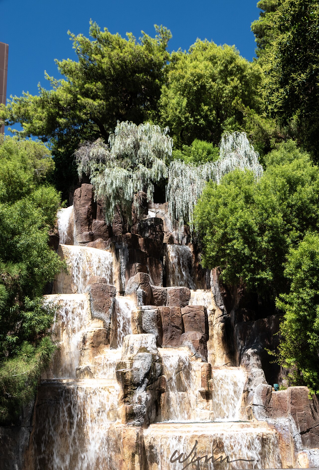 Terraced rock waterfall beneath mature pine trees outside Wynn Las Vegas, with the Wynn name on the lower basin.