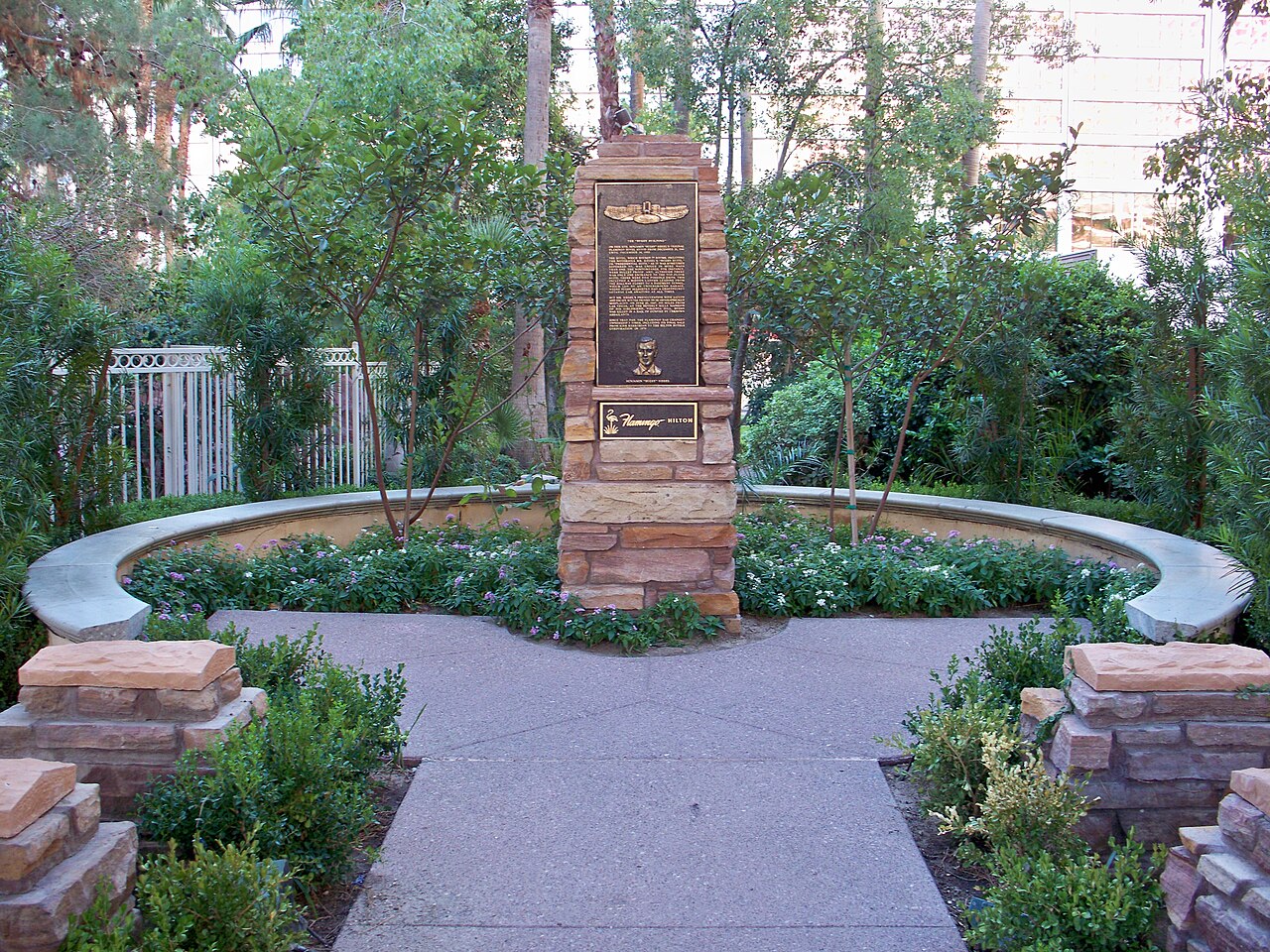 A bronze memorial plaque on a stone pillar stands in a circular garden seating area surrounded by shrubs.