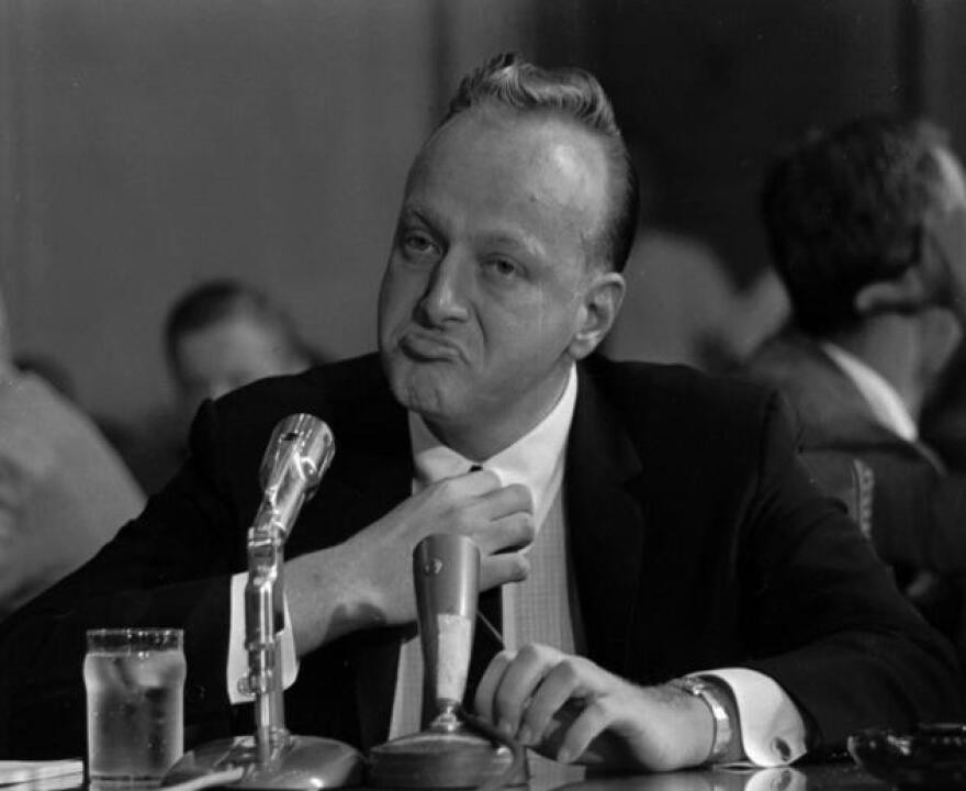 A black-and-white photograph of a suited man seated before microphones.