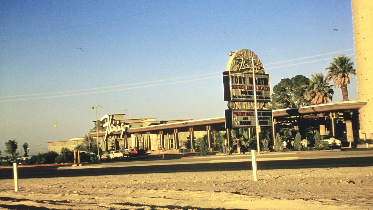 A low modern hotel frontage and freestanding Flamingo marquee face the road, with palm trees and parked cars.