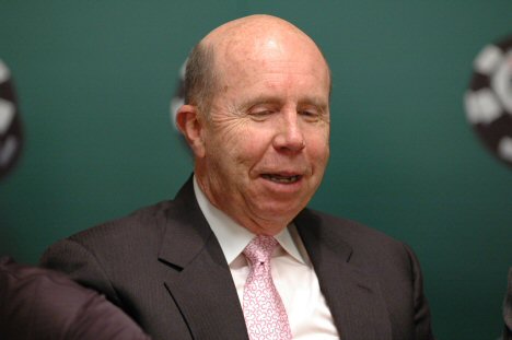 Jack Binion wearing a dark suit and pink tie, in front of a green backdrop.