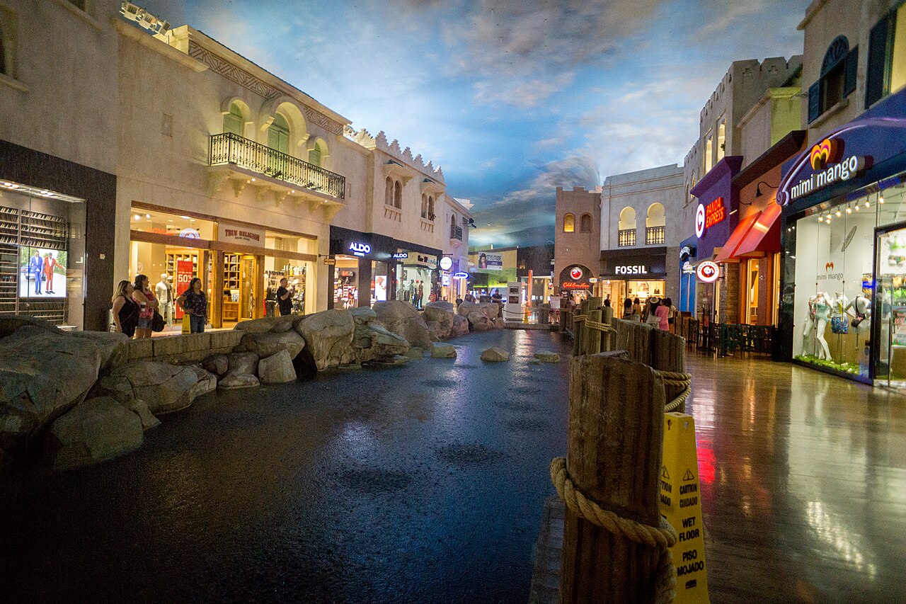 Rain splashes into a rock-lined indoor channel beneath a painted sky, with shopfronts on either side.