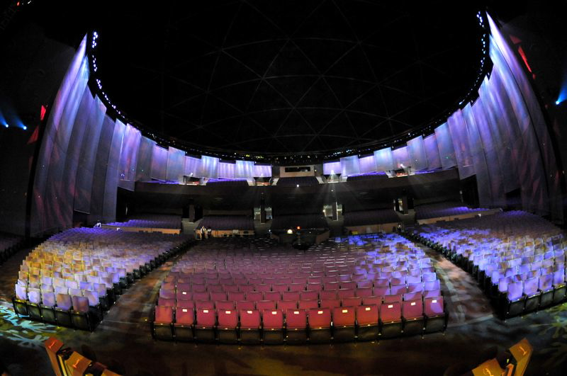 Rows of red and yellow seats beneath a dark domed roof and violet-lit walls, seen from the stage.