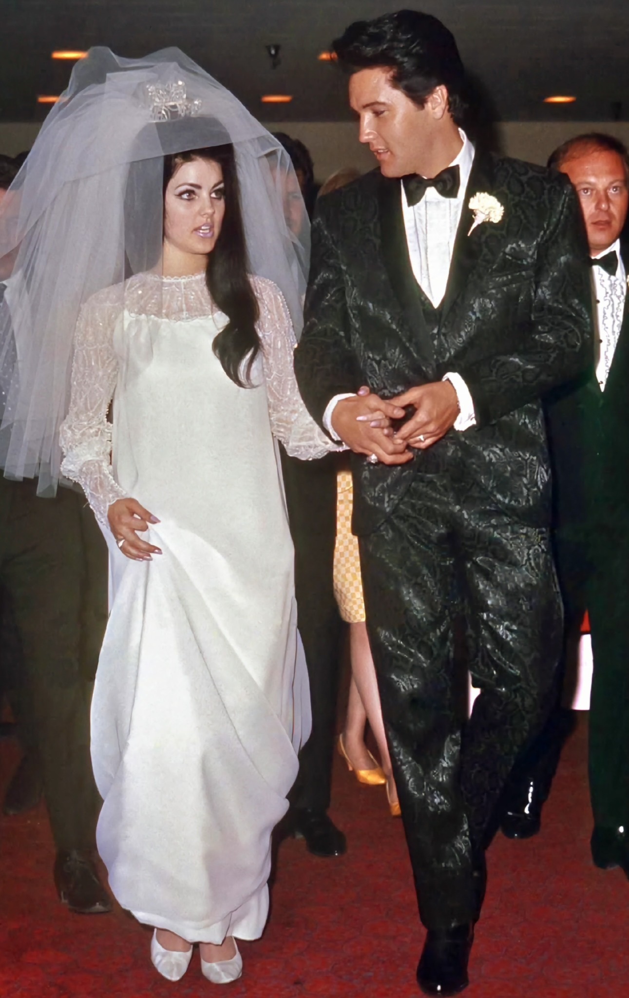 Priscilla in a white wedding dress and veil stands beside Elvis in a dark suit, their hands joined.