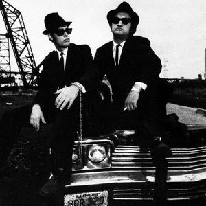 Two suited figures in hats and sunglasses seated on a car.