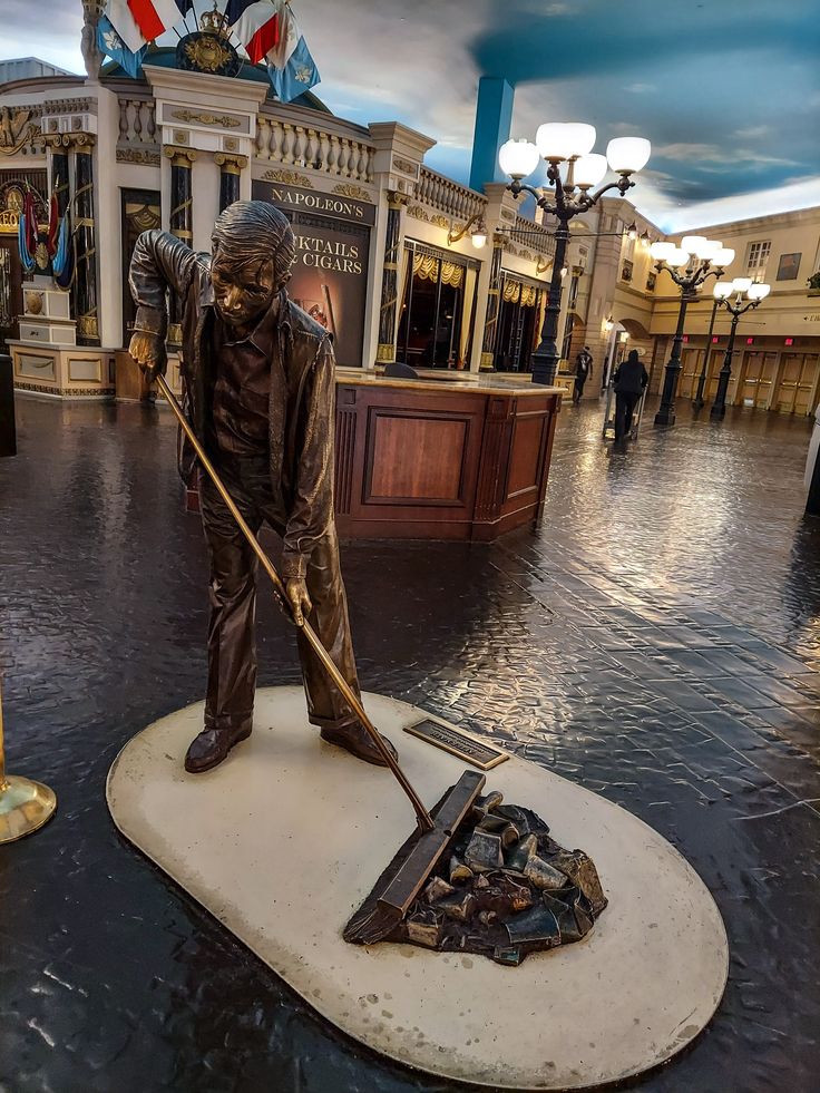 A bronze-coloured sweeper sculpture holding a broom beside a pile of debris in the Paris Las Vegas interior.