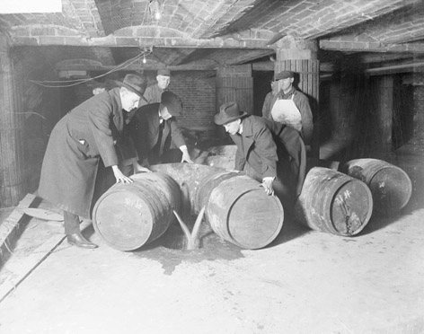 Men in coats and hats lean over wooden barrels while liquid spills across a cellar floor.