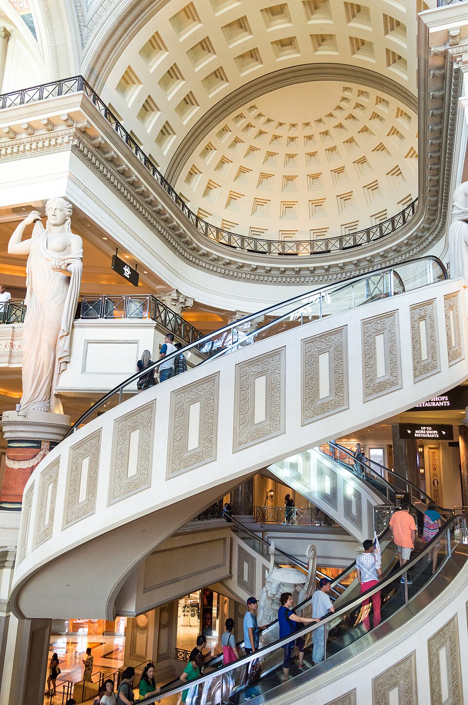 Curving escalators carry shoppers between floors beneath a domed ceiling in the Forum Shops, beside classical statues.