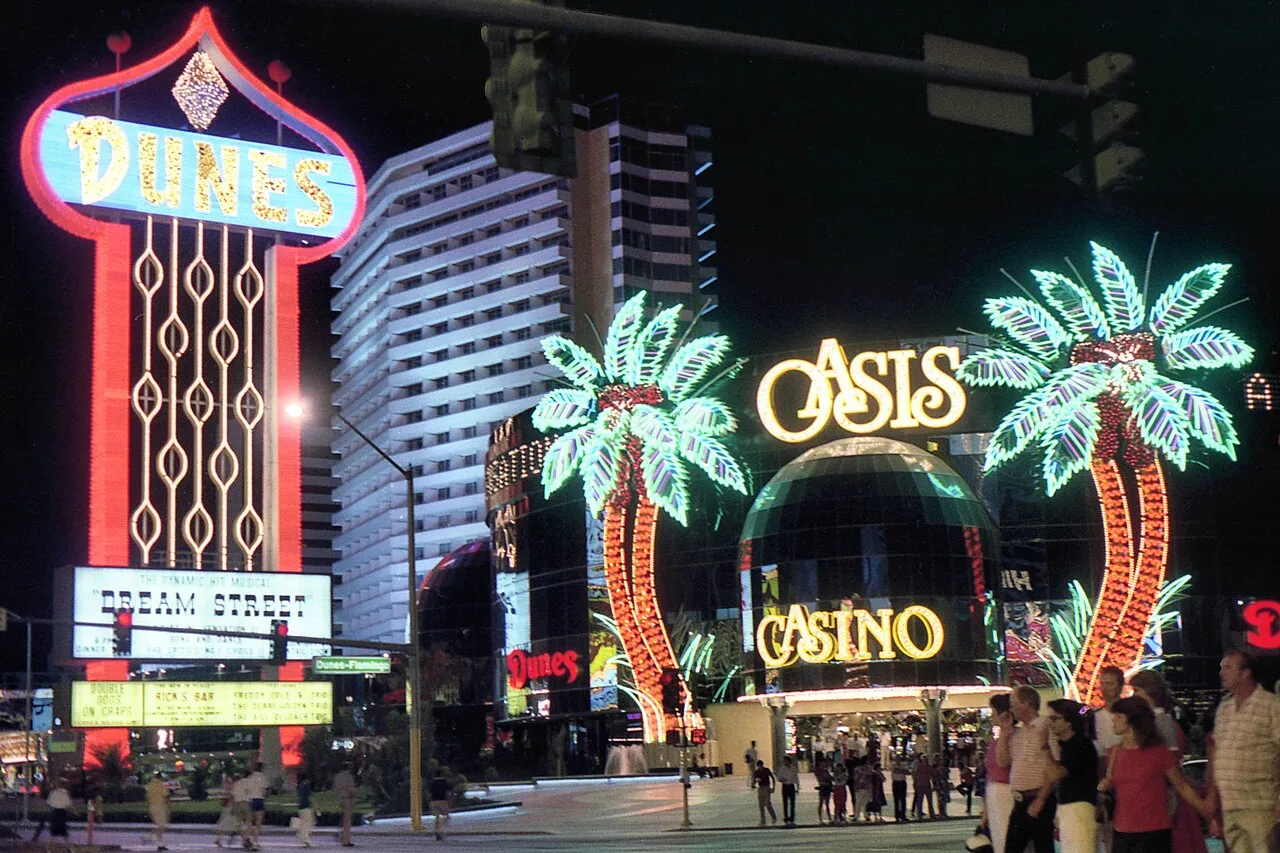 Illuminated Dunes pylon and hotel tower beside Oasis Casino’s round frontage and neon palm trees at night.