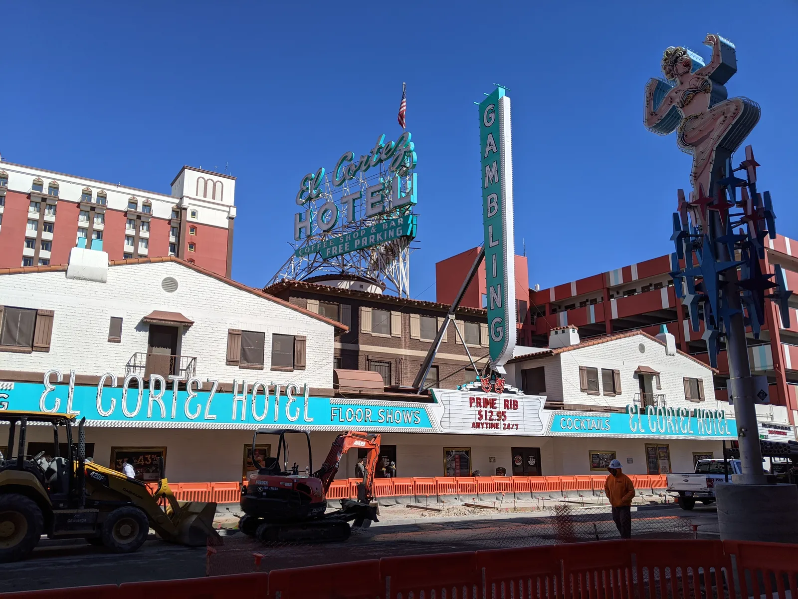 El Cortez’s white Fremont Street buildings beneath turquoise hotel and gambling signs, with its taller hotel tower behind and orange road-work barriers in front.