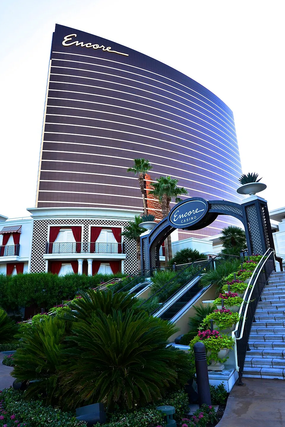 Encore’s curved bronze hotel tower above a casino entrance arch, landscaped steps, escalators and palm trees.
