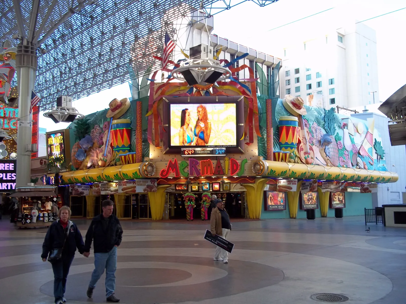 Mermaids’ colorful casino sign beneath a video screen, flanked by decorative drums, hats and mermaid figures, with pedestrians under the Fremont Street canopy.