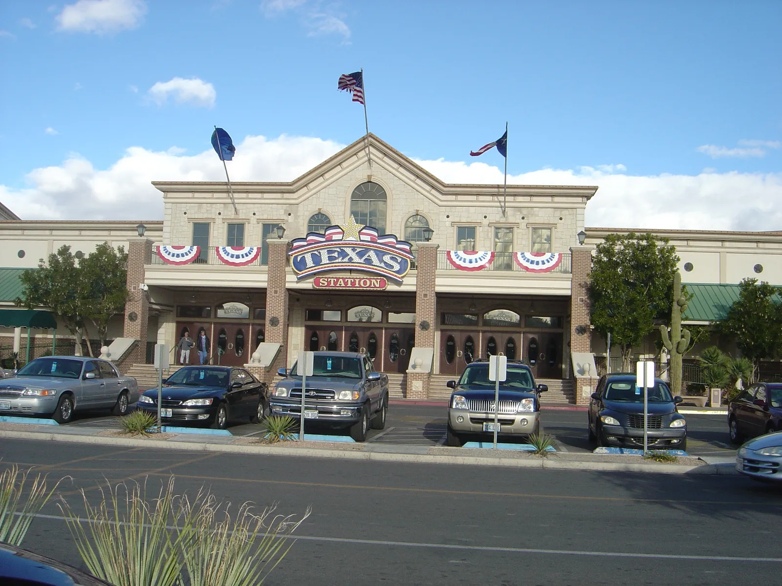 Texas Station’s cream and brick entrance facade with its Texas sign, flags and parked cars in front.
