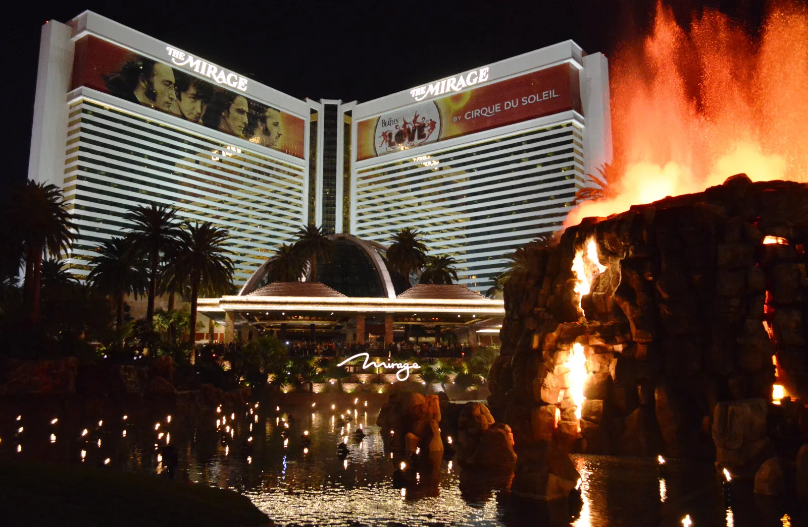 Fire erupts from the Mirage’s artificial volcano beside a lagoon, with the hotel’s illuminated entrance and tower carrying Beatles LOVE advertising behind it.