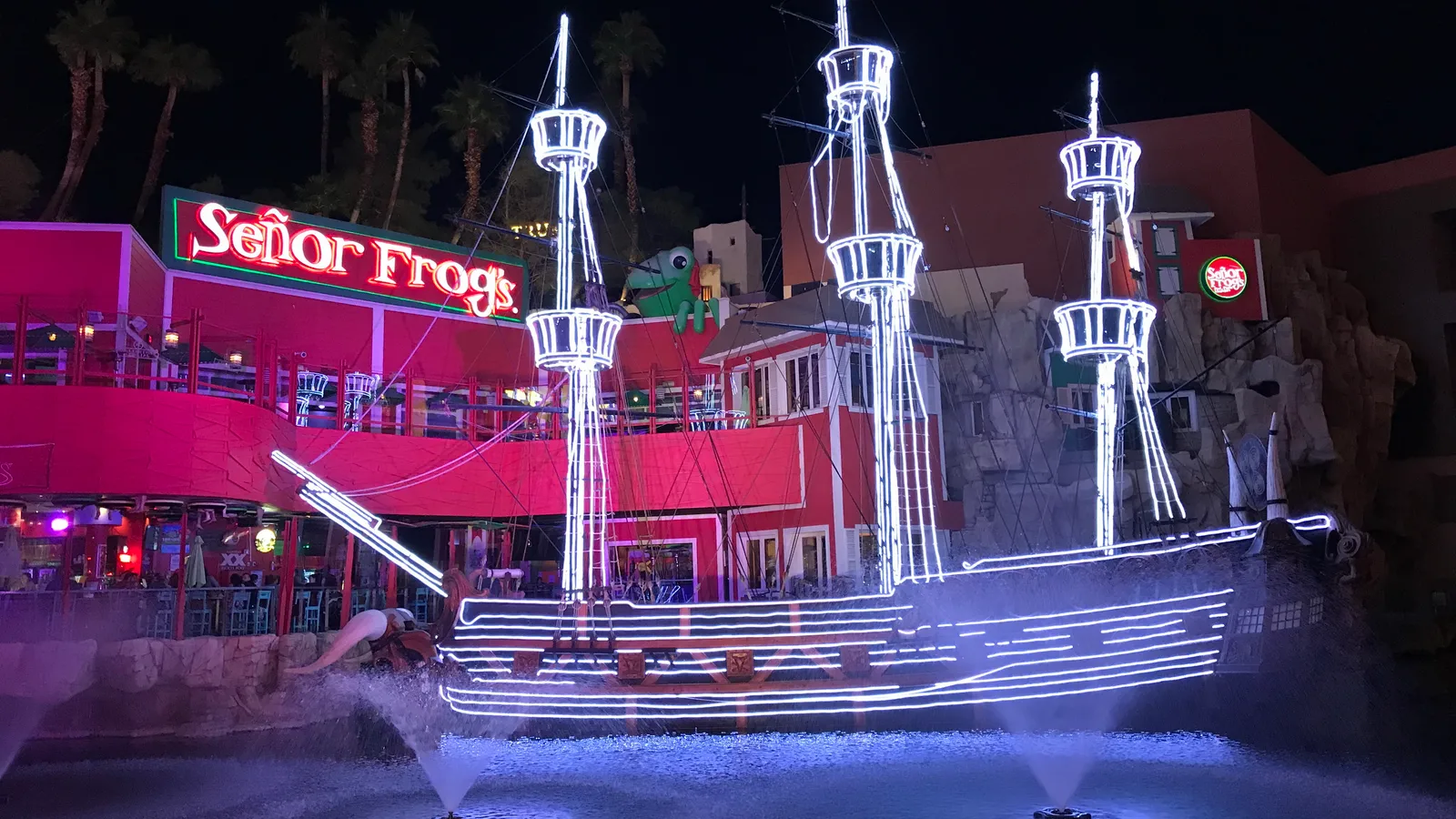 A retained sailing ship outlined in white lights in Treasure Island’s lagoon, with fountains and Señor Frog’s signs behind it; no live show is taking place.