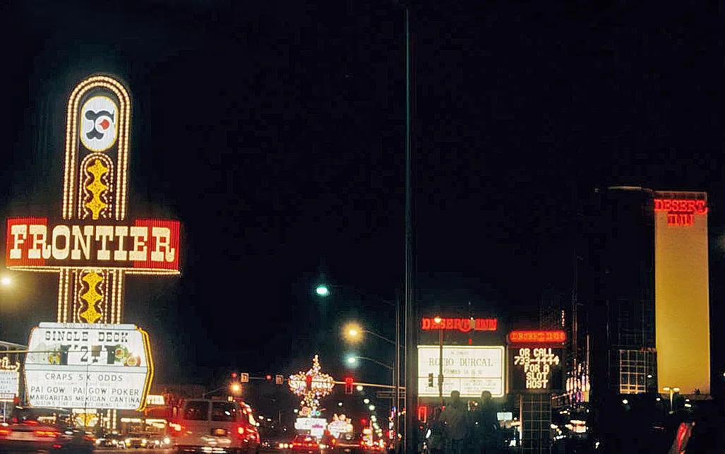 Nighttime Strip view with the Frontier sign at left and Desert Inn signs and a hotel tower at right.